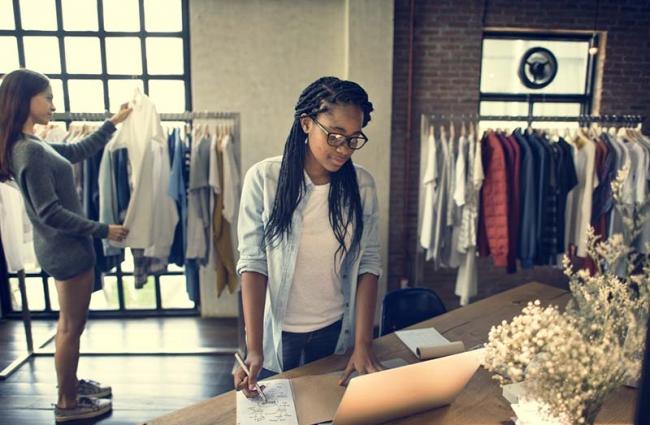 woman at laptop with another woman in background looking at clothes on a rack