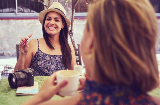 Woman vaping with e-cigarette at a table with another woman drinking coffee.