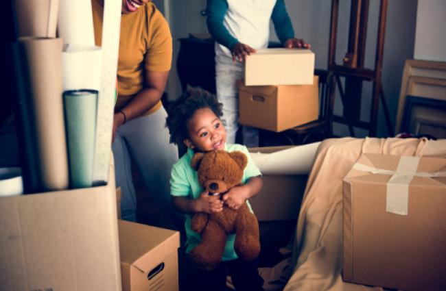 A smiling little girl holding a teddy bear while parents move boxes