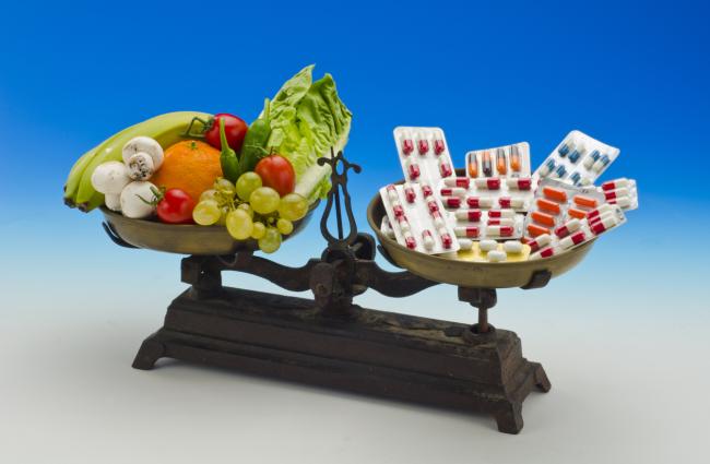 Image of a two-armed scale with vegetables and fruits on the left hand side and colorful pills on the right hand side.