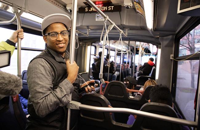Black man wearing a white cap, glasses, black vest, and charcoal shirt smiling while riding the bus