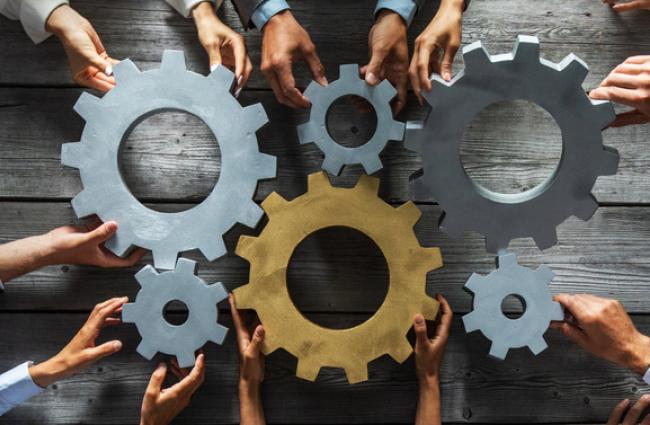 Group of people joining together silver and golden colored gears on table at workplace top view