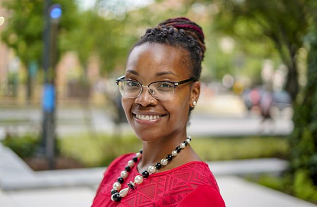 Dr. Jeana Morrison, a smiling Black woman with braided hair twisted into a bun at the back of her head. She's wearing glasses, a beaded necklace, and a red patterned top. She's standing in front of a blurred background of what looks like a park or part of a college campus.