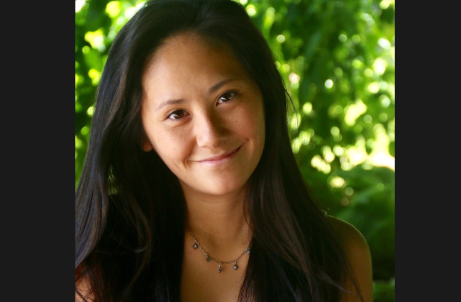 Elissa Carey, a smiling Chinese American woman with long dark hair. She's wearing a dangly beaded necklace and white top. She's sitting in front of a green leafy background.