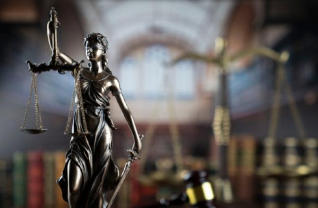 A brass-colored metal female statue holding up a balancing scale with her right arm, and in her left arm she is holding a long sword pointed toward the ground. The background is a blurred out library with a book shelf.