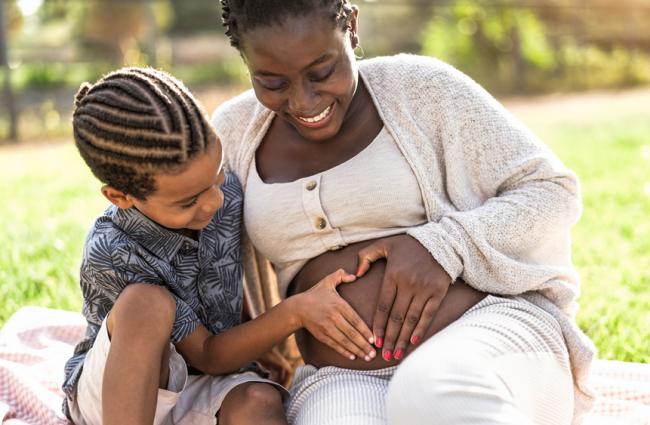 Black pregnant person sitting in a grassy area with a young Black child, both of whom have their hands on the pregnant person's abdomen