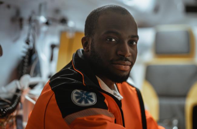 In the foreground, there’s a young Black man wearing an emergency medical response orange jacket and looking into the camera, and a blurred background of the inside of an ambulance.