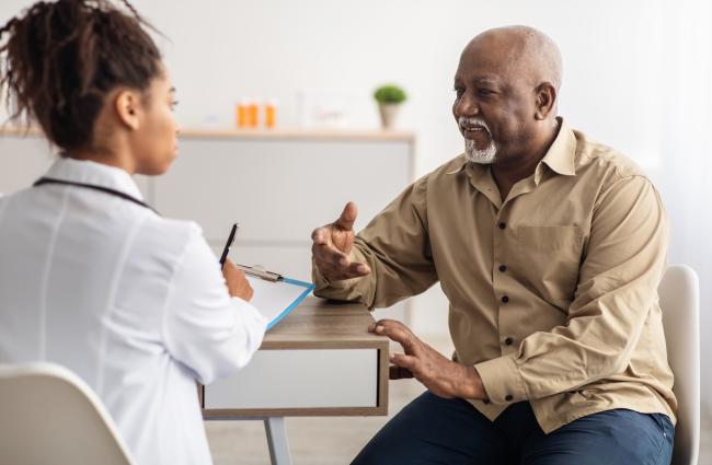 Black senior man sitting down and talking to a Black female doctor who is filling form listening to elderly patient