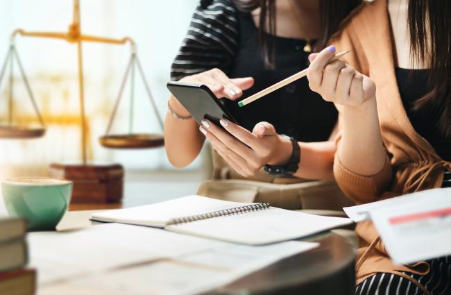 Two female-appearing people with manicured nails are looking at a phone over a desk. Underneath there are white notepads, and in the background is an old fashioned balance/weigh scale, insinuating some sort of law office.