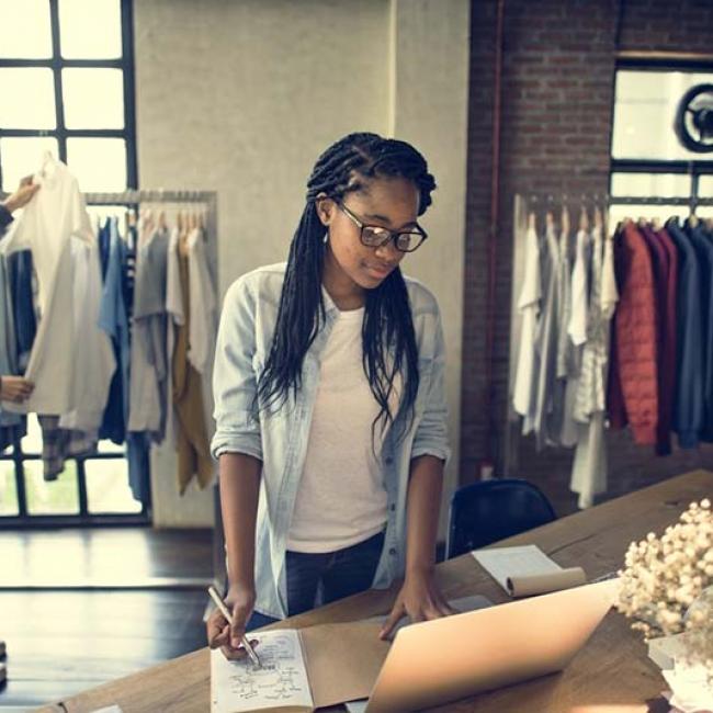 woman at laptop with another woman in background looking at clothes on a rack