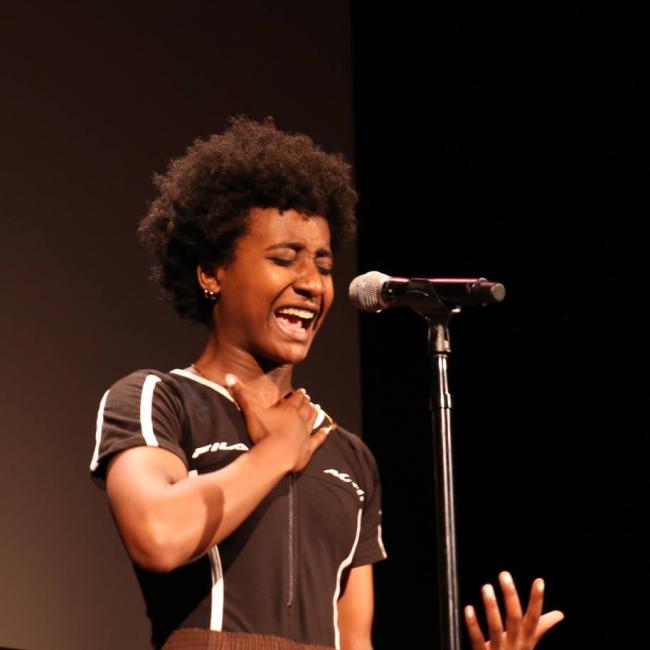 Young African American woman passionately speaking into a microphone on stage.