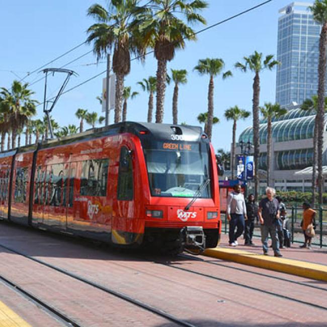 Red San Deigo light rail train at a stop on a sunny day and palm trees.