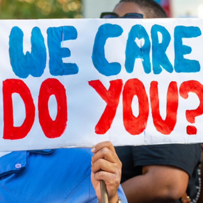 Image of a masked woman holding a sign with text: We care, do you?