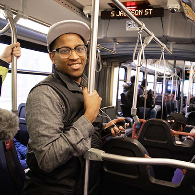 Black man wearing a white cap, glasses, black vest, and charcoal shirt smiling while riding the bus