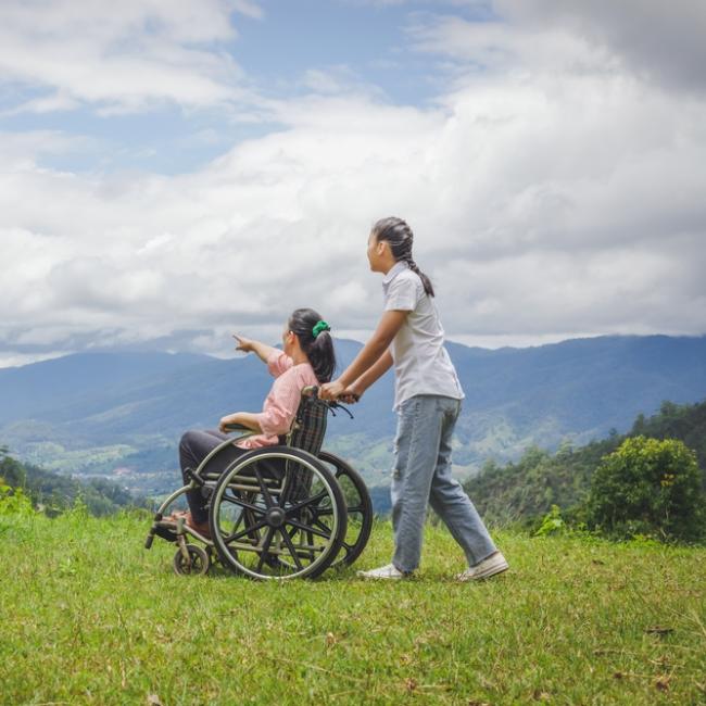 Woman in wheelchair and friend walking on hillside park on a sunny day