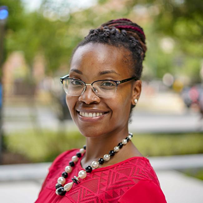 Dr. Jeana Morrison, a smiling Black woman with braided hair twisted into a bun at the back of her head. She's wearing glasses, a beaded necklace, and a red patterned top. She's standing in front of a blurred background of what looks like a park or part of a college campus.