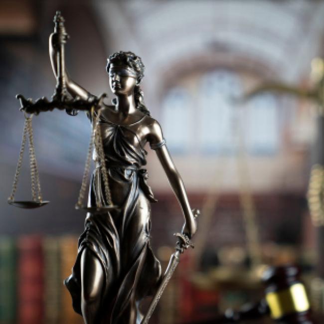 A brass-colored metal female statue holding up a balancing scale with her right arm, and in her left arm she is holding a long sword pointed toward the ground. The background is a blurred out library with a book shelf.