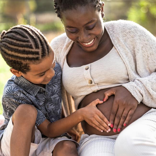 Black pregnant person sitting in a grassy area with a young Black child, both of whom have their hands on the pregnant person's abdomen
