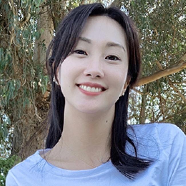 Dr. Ye Ji Kim, a dark-haired Korean American female-identifying epidemiologist wearing a light blue top and standing in front of a tree