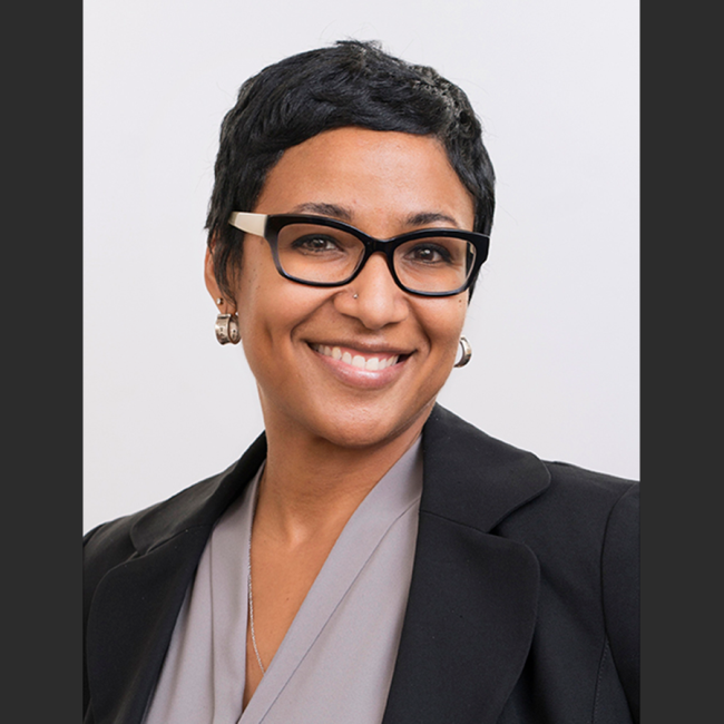 Dr. Amani Allen, a smiling Black woman with short black hair, wearing black glasses a grey blouse, and a dark blazer