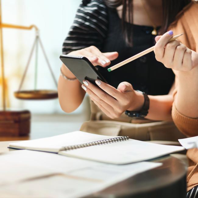 Two female-appearing people with manicured nails are looking at a phone over a desk. Underneath there are white notepads, and in the background is an old fashioned balance/weigh scale, insinuating some sort of law office.