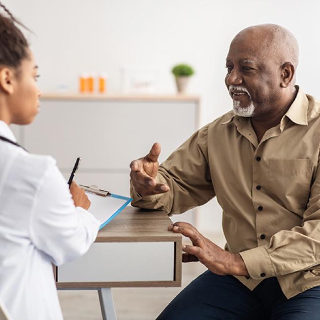 Black senior man sitting down and talking to a Black female doctor who is filling out a form while listening to an elderly patient