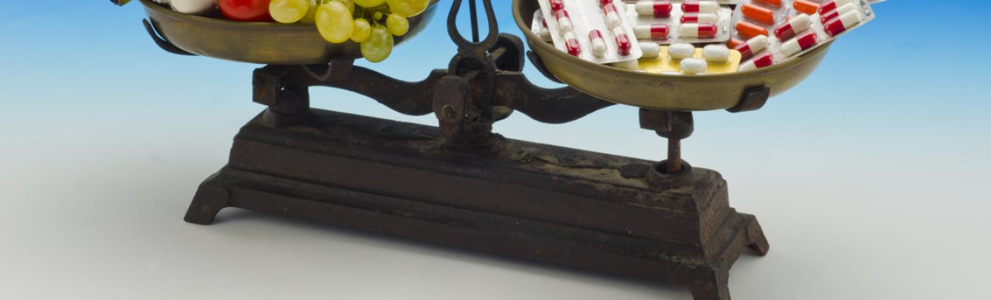 Image of a two-armed scale with vegetables and fruits on the left hand side and colorful pills on the right hand side.