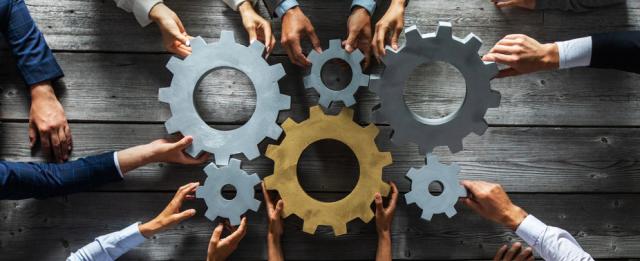 Group of people joining together silver and golden colored gears on table at workplace top view