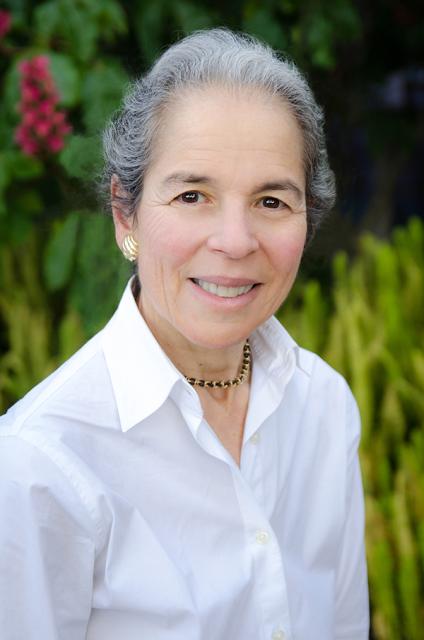 Nancy Adler, a smiling white woman with silver and gray hair wearing gold earrings and a white shirt