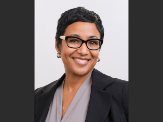 Dr. Amani Allen, a smiling Black woman with short black hair, wearing black glasses a grey blouse, and a dark blazer